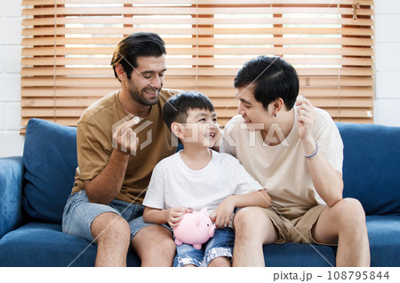 Happy LGBTQ+ gay family with adopted little son stay together in living room and teaching their adopted son about wealth and saving money for future. Diversity in gender and lifestyle concept. Happy LGBTQ+ gay family with adopted little son stay together in living room and teaching their adopted son about wealth and saving money for future. Diversity in gender and lifestyle concept. 108795844