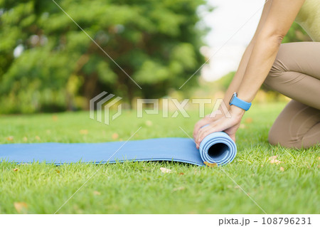 Happy beautiful Asian woman rolling out an exercise - yoga mat on the grass field to prepare doing a yoga and mediation exercise. Woman preparing a yoga or body weight and meditating exercise. Happy beautiful Asian woman rolling out an exercise - yoga mat on the grass field to prepare doing a yoga and mediation exercise. Woman preparing a yoga or body weight and meditating exercise. 108796231