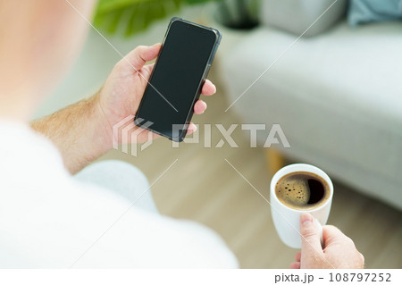 Caucasian white man wake up from the bed in morning and sitting beside the bed and holding an empty blank screen smartphone and enjoy drinking his morning coffee. Man checking message on smartphone. 108797252