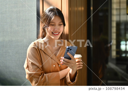 Happy young woman with take away coffee cup using mobile phone while standing near office building 108798434