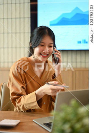 Cheerful Asian female employee looking at laptop screen and talking on mobil phone Cheerful Asian female employee looking at laptop screen and talking on mobil phone 108798508