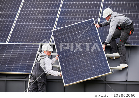 Builders building solar panel system on roof of house. Men workers in helmets carrying photovoltaic solar module outdoors. Concept of alternative and renewable energy. 108802060