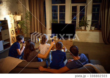 Back view. Family, parents and children sitting on couch in evening at home and watching football match translation Back view. Family, parents and children sitting on couch in evening at home and watching football match translation 108802503