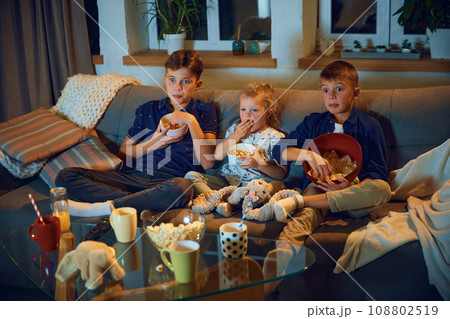 Children, brothers and little sister sitting on couch in living room in evening, watching tv together and eating snacks. Children, brothers and little sister sitting on couch in living room in evening, watching tv together and eating snacks. 108802519