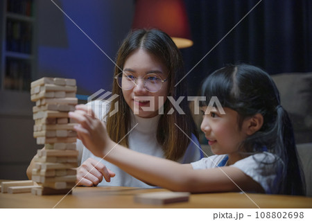 Asian young mother playing game in wood block with little daughter in home living room at night, Smiling woman help teach preschooler kid play build constructor tower of wooden blocks, family funny Asian young mother playing game in wood block with little daughter in home living room at night, Smiling woman help teach preschooler kid play build constructor tower of wooden blocks, family funny 108802698