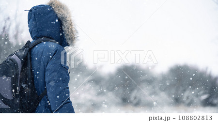 Back view of traveler in a blue jacket with a fur hood and a backpack on the background of a winter landscape during snowfall. 108802813