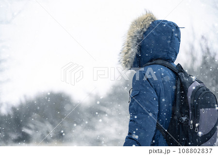 Back view of traveler in a blue jacket with a fur hood and a backpack on the background of a winter landscape during snowfall. 108802837