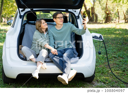Loving couple with smartphones sits in an electric car's trunk 108804097
