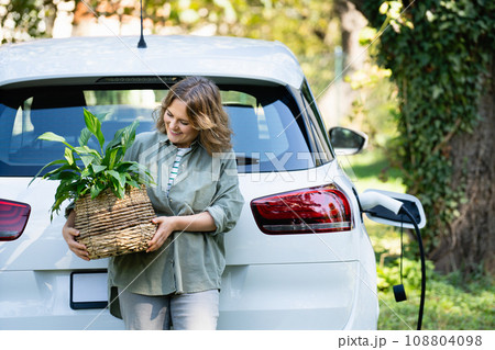 Woman with plant in pot next to a charging electric car in the yard of a country house Woman with plant in pot next to a charging electric car in the yard of a country house 108804098