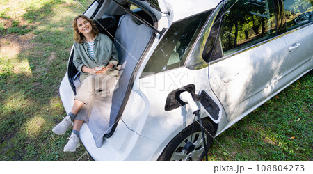 Woman with smartphone sits in an electric car's trunk 108804273