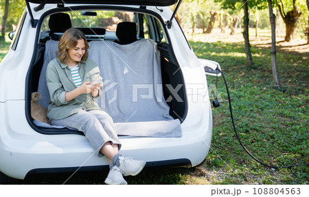 Woman with smartphone sits in an electric car's trunk Woman with smartphone sits in an electric car's trunk 108804563