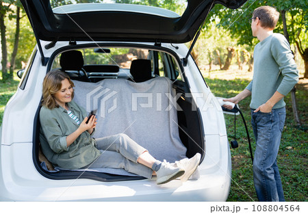 Woman with smartphone sits in an electric car's trunk when the man holds electric plug Woman with smartphone sits in an electric car's trunk when the man holds electric plug 108804564