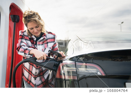 Woman in a plaid shirt charging an electric car 108804764