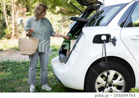 Woman with shopping bag next to a charging electric car in the yard of a country house Woman with shopping bag next to a charging electric car in the yard of a country house 108804768