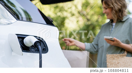 Woman with shopping bag next to a charging electric car in the yard of a country house Woman with shopping bag next to a charging electric car in the yard of a country house 108804811