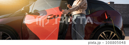 Young woman is standing near the electric car and holding smartphone Young woman is standing near the electric car and holding smartphone 108805048