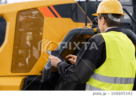 Worker in a helmet with a digital tablet on the background of construction machine Worker in a helmet with a digital tablet on the background of construction machine 108805341