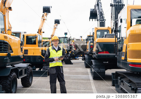 Engineer in a helmet with a digital tablet stands next to construction excavators. 108805387