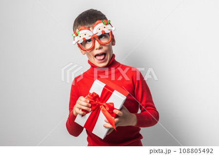 Funny grimacing child boy wearing fun glasses in form of Christmas Santa Claus holding Xmas gift while standing in a white background, dressed in warm red xmas sweater. Generation alpha and gen alpha 108805492