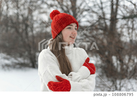 Beautiful girl in warm knitted sweater, red hat and mittens poses in nature in snowy park. Woman in knitwear outdoors 108806795