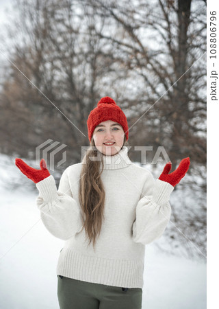 Portrait of beautiful young woman in woolen hat outdoors, cold weather. Young woman in knitted red hat and mittens, winter park. Vertical frame 108806796