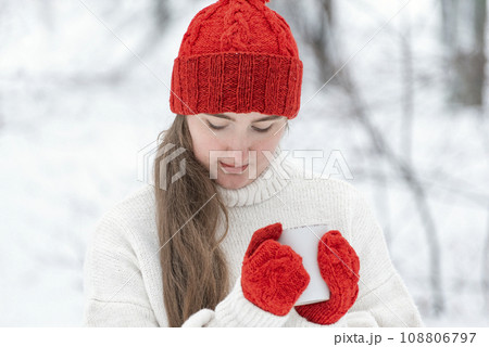 Beautiful young woman drink hot beverage from cup in winter outdoor. Portrait of pensive young woman in red hat and mittens in snowy park 108806797