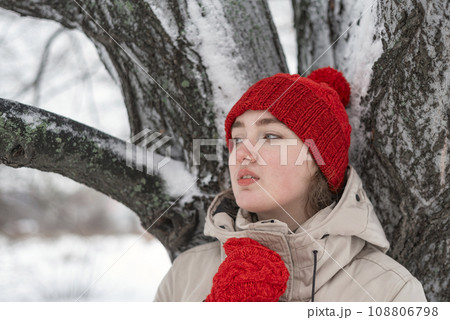Portrait of beautiful girl thoughtfully looking into distance. Young woman in red knitted hat and mittens stands near snow-covered tree. 108806798