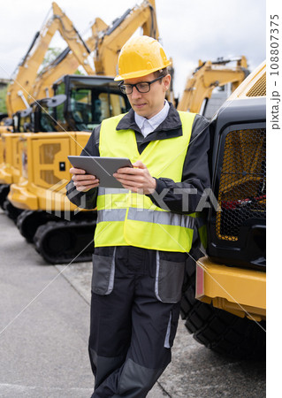 Engineer in a helmet with a digital tablet stands next to construction excavators. 108807375
