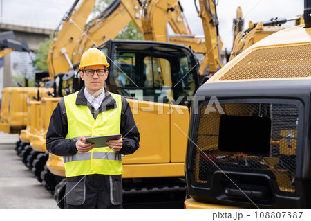Engineer in a helmet with a digital tablet stands next to construction excavators. 108807387