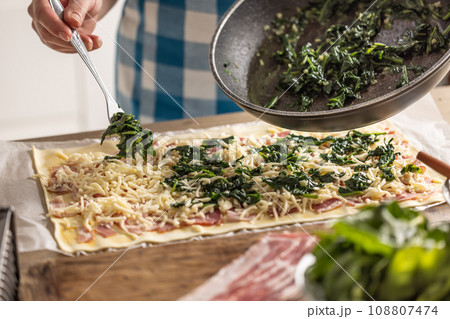 The cook prepares mini pizza cakes, puts spinach on puff pastry with cheese and bacon. The cook prepares mini pizza cakes, puts spinach on puff pastry with cheese and bacon. 108807474