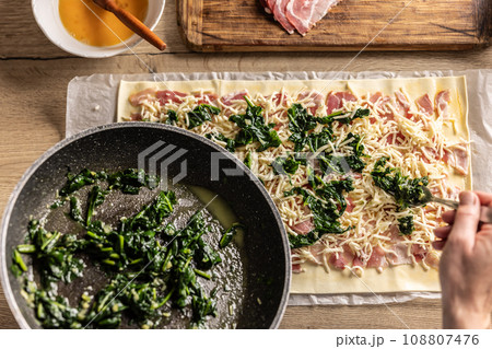 The cook prepares mini pizza cakes, puts spinach on puff pastry with cheese and bacon. The cook prepares mini pizza cakes, puts spinach on puff pastry with cheese and bacon. 108807476