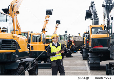 Engineer in a helmet with a digital tablet stands next to construction excavators. 108807553