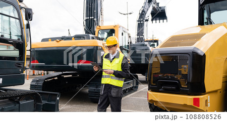 Engineer in a helmet with a digital tablet stands next to construction excavators Engineer in a helmet with a digital tablet stands next to construction excavators 108808526