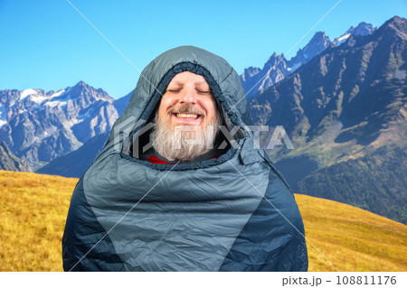 Happy bearded man in a sleeping bag against the backdrop of nature in the mountains. Happy bearded man in a sleeping bag against the backdrop of nature in the mountains. 108811176