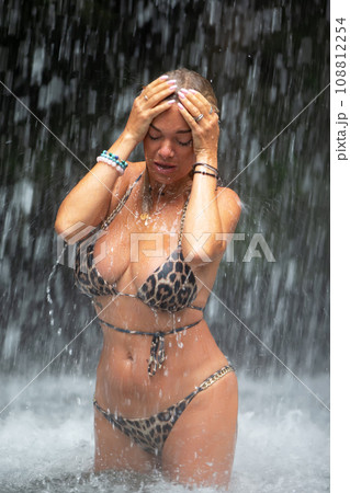 Beautiful young woman relaxes under a waterfall. Sexy girl in a bikini posing near a waterfall in the tropics. A traveler bathes under streams of water flowing from the mountains. 108812254