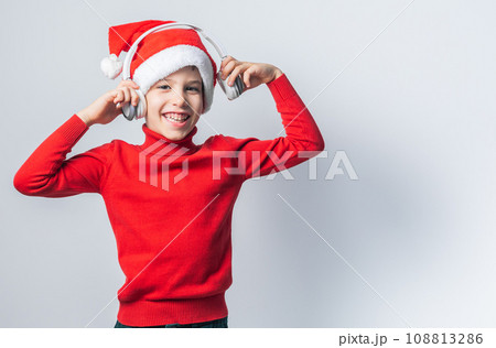 Funny child boy holds listening to Christmas music with Santa Claus hat on white background with copy space. Generation alpha and gen alpha children. 108813286