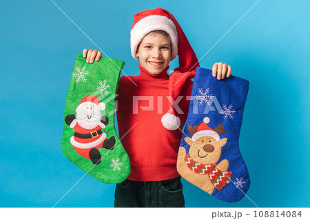 Child boy in red sweater and Santa hat with Christmas socks on blue background with space for text Child boy in red sweater and Santa hat with Christmas socks on blue background with space for text 108814084