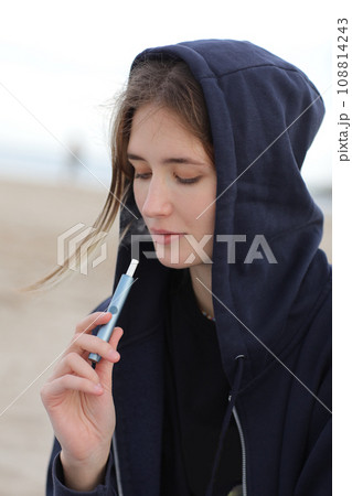 Electronic cigarette technology. Young woman smokes and releases steam from hybrid cigarette device that uses real tobacco refills with a heating pad, tobacco heating system. Bad unhealthy habit Electronic cigarette technology. Young woman smokes and releases steam from hybrid cigarette device that uses real tobacco refills with a heating pad, tobacco heating system. Bad unhealthy habit 108814243