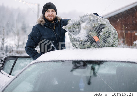 Handsome man tying to put a Christmas tree to the roof of the car to bring it home. Live fir tree devilery 108815323
