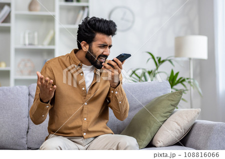Angry angry man talking on the phone at home, dissatisfied customer sitting on the sofa in the living room yelling at the interlocutor. Angry angry man talking on the phone at home, dissatisfied customer sitting on the sofa in the living room yelling at the interlocutor. 108816066