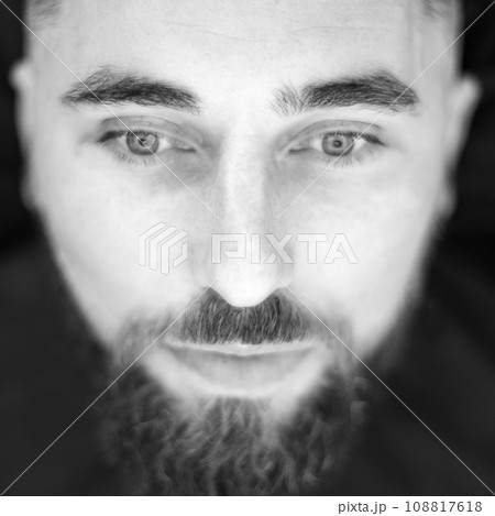 Black and white portrait of a bearded man closeup. A young man with a beard in a black cape sits on a chair in a barbershop. The client is waiting for a haircut of the beard. 108817618