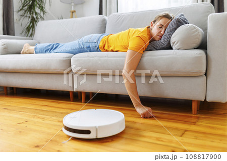 A woman sleeping on sofa while robot vacuum quietly takes care of the cleaning 108817900