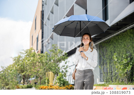 In the sweltering weather, a young businesswoman holds an umbrella while walking to the office. Her stern expression and successful demeanor are evident despite the hot temperature. 108819073