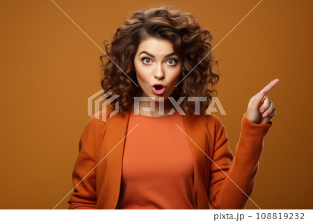 Excited woman pointing finger to side, shocked female person isolated on studio background. Portrait of surprised adult girl against brown wall. Concept of show, shopping, shock, sale 108819232