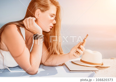 Digital nomad, woman in the hat, a business woman with a laptop sits on the rocks by the sea during sunset, makes a business transaction online from a distance. Freelance, remote work on vacation. 108820373