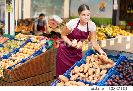 Girl seller of fruit and vegetable store works near showcase with garden-stuff, sorting yam Girl seller of fruit and vegetable store works near showcase with garden-stuff, sorting yam 108821833