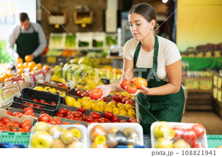 Girl seller of garden stuff store works near showcase, sorting tomato, checking goods 108821941