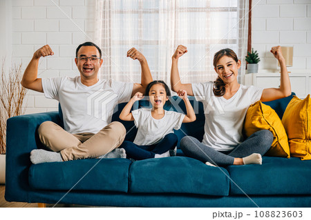 In their cozy living room a family day concept unfolds as young parents their child and daughter highlight their biceps muscles showcasing strength and togetherness with smiles and joy. In their cozy living room a family day concept unfolds as young parents their child and daughter highlight their biceps muscles showcasing strength and togetherness with smiles and joy. 108823603