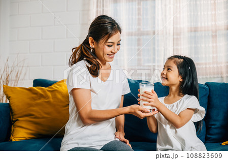 A loving Asian mother shares a glass of milk with her daughter on the sofa creating a heartwarming picture of bonding growth and the essential role of calcium in their daily life. A loving Asian mother shares a glass of milk with her daughter on the sofa creating a heartwarming picture of bonding growth and the essential role of calcium in their daily life. 108823609
