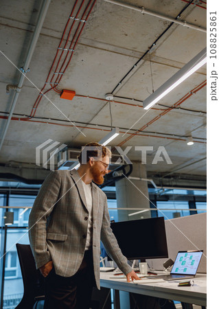 Smiling entrepreneur working with documents standing near desk in modern office Smiling entrepreneur working with documents standing near desk in modern office 108825861